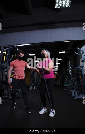 Verticale full length shot di una donna anziana e la sua fintess allenatore indossare maschere mediche al viso in palestra Foto Stock