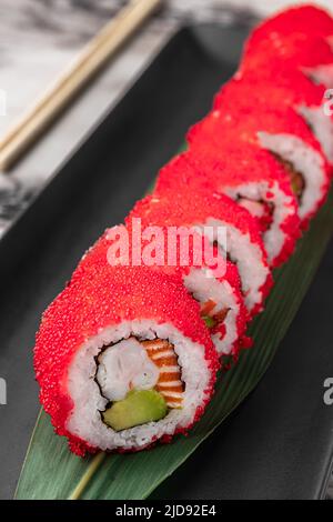 set di rotoli di philadelphia con caviale di pesce volante rosso, avocado, salmone e foglia di bambù verde in un piatto di ceramica nera con bacchette su un textu luminoso Foto Stock