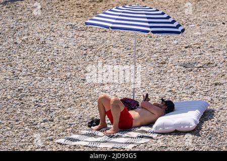 Monaco di Baviera, Germania. 19th giugno 2022. Un uomo si trova sotto il suo ombrellone su una sponda di ghiaia vicino al fiume Isar. Credit: Peter Kneffel/dpa/Alamy Live News Foto Stock