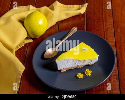 Una fetta di cheesecake al limone su un piatto scuro con un cucchiaino d'oro e decorazione floreale Foto Stock