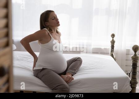 La donna incinta si siede sul letto toccherà la sua parte inferiore dolorante della schiena Foto Stock