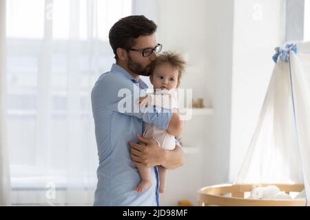 Daddy bacia il bambino in piedi vicino al presepe moderno in una stanza dei bambini accogliente Foto Stock