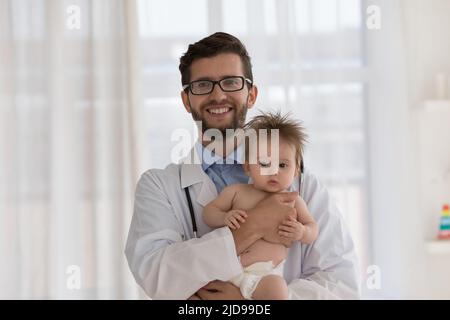 Pediatra in uniforme bianca che tiene il bambino cute guarda la macchina fotografica Foto Stock