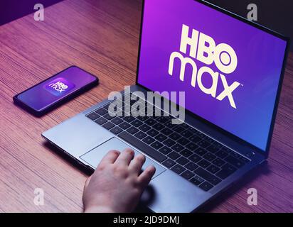 Giovane donna che guarda HBO Max su un portatile. Logo HBO Max sugli schermi di computer portatili e smartphone. Scrivania in legno con computer portatile e elegante Foto Stock