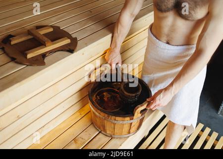 Uomo che raccoglie acqua da secchio di legno in sauna. Il centro benessere è disponibile nel bagno turco Foto Stock