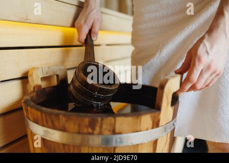 Primo piano di un uomo in un asciugamano che raccoglie acqua con un mestolo da un secchio di legno in una sauna Foto Stock