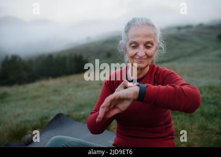 Una donna anziana jogger impostazione e guardare lo smartwatch sportivo, controllando le sue prestazioni in natura al mattino presto con nebbia e montagne in backgrou Foto Stock