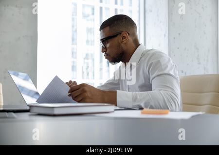 Serio uomo d'affari africano che lavora con documenti utilizzando un notebook cercando concentrato Foto Stock