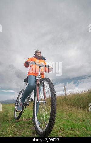 Giovane ispanica vista da sotto indossando giacca arancione e pantaloni blu cavalcando la sua bicicletta da sola attraverso la campagna rurale durante una giornata nuvolosa Foto Stock