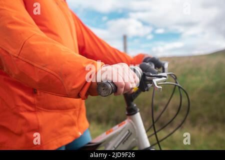 Dettaglio delle mani di una giovane donna che indossa una giacca arancione sul manubrio della bicicletta in mezzo al campo in una giornata nuvolosa Foto Stock