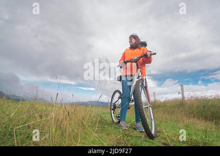 Giovane ispanica vista da sotto indossando una giacca arancione e pantaloni blu che riposano sulla sua bicicletta nel mezzo di un campo rurale durante una giornata nuvolosa Foto Stock