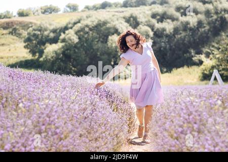 La ragazza di fuoco selettivo morbido in abito viola cammina sul campo di lavanda e gode l'aroma di fiori rosa e la bellezza in giorno di sole. Colore trendy di 2022 molto peri. Detox digitale piacere della vita lenta. Foto Stock