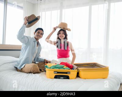 Coppia di viaggiatori che si preparano a partire. Giovane donna asiatica e uomo in denim camicia con cappelli da spiaggia mentre si impacchettano con valigie gialle sul letto nel letto Foto Stock