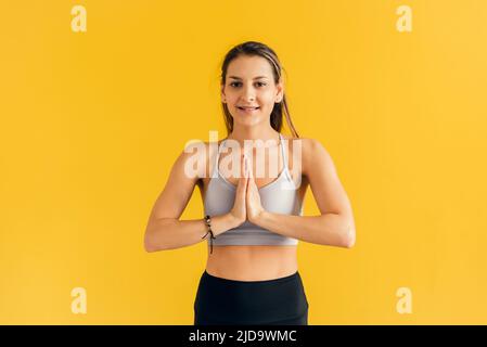 Giovane donna concentrata in abbigliamento sportivo stretto che pratica yoga, tenendo le mani nel gesto namaste e meditando, rilassandosi, guardando la macchina fotografica. Donna Foto Stock