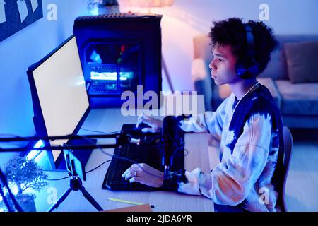 Vista laterale ritratto di ragazzo teenage nero giocare video gioco su PC e streaming dal vivo in blu luce neon Foto Stock