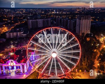 Vista ravvicinata della ruota panoramica illuminata dall'antenna. Zona ricreativa del centro di Kharkiv con luci serali. Divertimento Gorky Central Park nei colori del tramonto Foto Stock