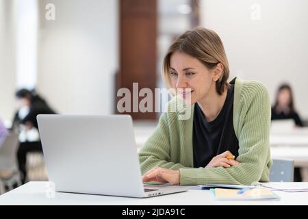 Sorridente donna di mezza età che ama lo studio online, guardando il webinar tramite notebook in biblioteca Foto Stock