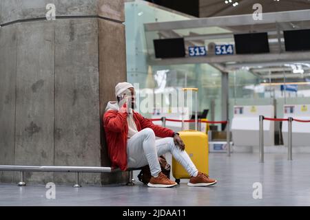 Gioioso bel maschio nero con giacca rossa sorridente facendo una chiamata al telefono cellulare mentre aspettate il volo Foto Stock