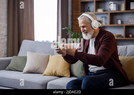Felice uomo anziano dai capelli grigi in cuffie bianche che tengono lo smartphone e ridono. Vecchio uomo seduto sul divano a casa guardando contenuti divertenti Foto Stock
