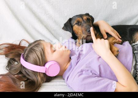 Ragazza felice adolescente in t-shirt lilla in cuffie rosa ascolta la musica e ha divertimento a giocare con il cane Foto Stock