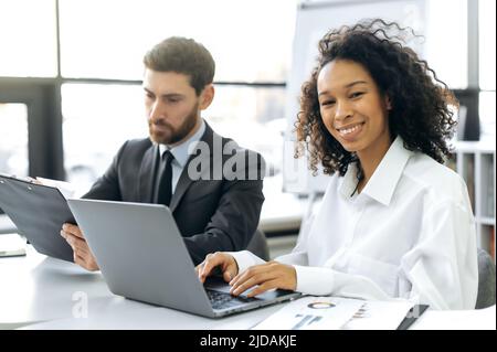 Colleghi internazionali che lavorano in un ufficio moderno, uomo caucasico, manager, che lavorano con i documenti, donna afroamericana, consulente finanziario, lavora a un computer portatile, guarda la fotocamera, sorridendo Foto Stock