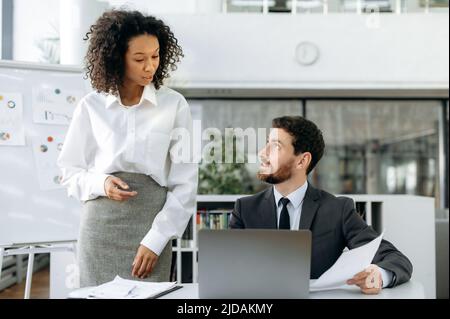 Colleghi di affari multirazziali, donna afroamericana e uomo caucasico, partner di successo che lavorano insieme in un ufficio moderno utilizzando un computer portatile, discutere un piano per una riunione, fare una presentazione Foto Stock