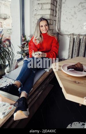 Donne piuttosto giovani che bevono caffè caldo in un caffè. Persona che tiene una tazza di cappuccino sopra la finestra mentre riposa in città. Ospite che ha un freno corto Foto Stock