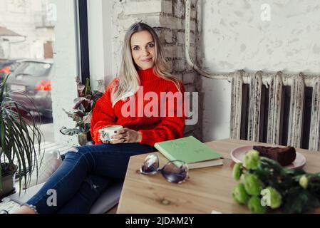 Donne piuttosto giovani che bevono caffè caldo in un caffè. Persona che tiene una tazza di cappuccino sopra la finestra mentre riposa in città. Ospite che ha un freno corto Foto Stock