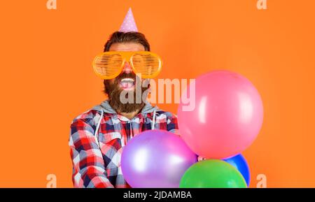 Ragazzo bearded in occhiali divertenti e cappello cono con palloncini. Uomo in festa. Buon compleanno festeggiamenti. Foto Stock