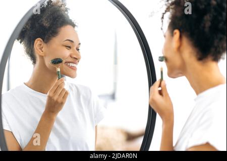 Cura della pelle del viso, prevenzione delle rughe. Bella giovane africana americana capelli ricci donna, facendo le procedure di cura della pelle del viso, in piedi davanti ad uno specchio a casa, usando un rullo di giada, sorridendo felicemente Foto Stock