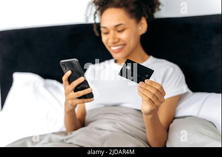 Smartphone e carta di credito in mani femminili. Fanciulla afroamericana defocused, sdraiata su un letto a casa, usando il cellulare e la carta di credito per lo shopping online, ordinando la consegna online, sorridendo. Tecnologia Foto Stock