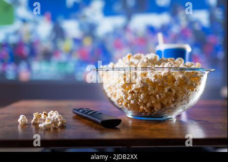 Su un tavolo di legno c'è una ciotola di popcorn, una bevanda in un bicchiere e un telecomando TV su uno sfondo sportivo. Guardare programmi sportivi in TV. Sport Foto Stock