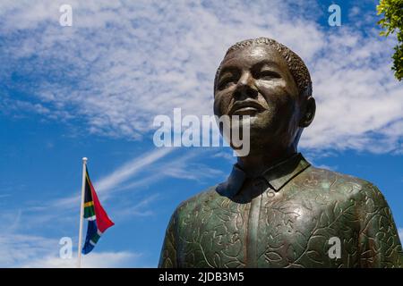 Primo piano della statua di bronzo Nelson Mandela in Piazza Noble lungo il Lungomare Victoria and Alfred con la bandiera sudafricana che vola contro un ... Foto Stock