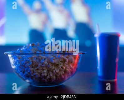 Guardare programmi musicali, film e serie in TV. Su un tavolo di legno vi sono popcorn in una ciotola trasparente di vetro e una bevanda analcolica in un vetro di plastica con una st Foto Stock