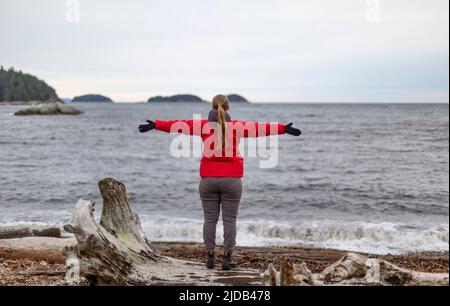 La donna si trova sul bordo dell'acqua e guarda l'oceano con le braccia allungate, Sergeant Bay, Sunshine Coast; British Columbia, Canada Foto Stock