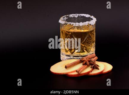 Un bicchiere di sidro cristallino con zucchero intorno al bordo, fette di mela rossa, anice stellato e bastoncini di cannella su sfondo nero. Primo piano. Foto Stock