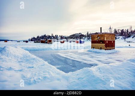 Vista su una pista di pattinaggio e su baracche e rimorchi per la pesca sul ghiaccio sulla costa del lago Frozen Windermere e del parco provinciale del lago Windermere Foto Stock