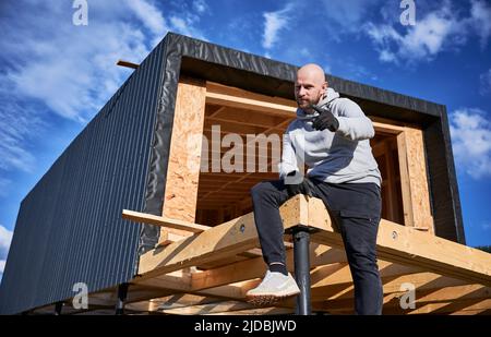 Maschio sviluppatore edificio legno telaio casa su palo fondazione. Ritratto di uomo calvo seduto sul terrazzo in cantiere, ispezionando la qualità del lavoro. Foto Stock