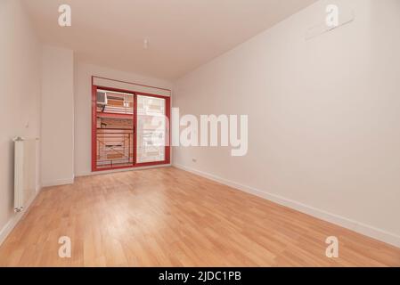 Camera vuota con pareti dipinte di bianco, pavimento in parquet di quercia, radiatore in alluminio bianco e Falegnameria in alluminio rossastro sulle porte scorrevoli verso il balcone Foto Stock