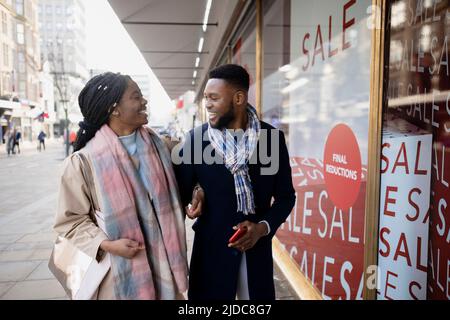 Allegro braccio di coppia in braccio camminando oltre i segni di vendita nella finestra del negozio in città Foto Stock