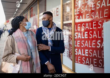 Coppia che indossa i rivestimenti del viso shopping, passando vetrina con segno di vendita Foto Stock