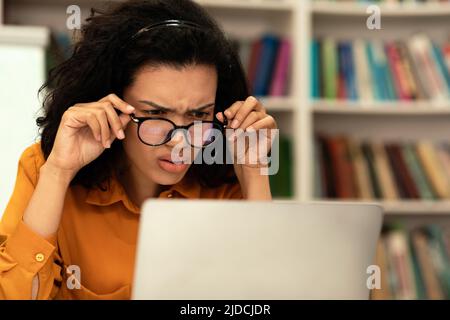 Donna da corsa mista concentrata in occhiali che squinting e guardando il portatile, seduto in biblioteca e studiare online Foto Stock