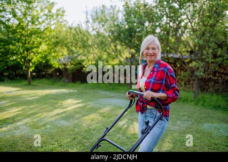 Donna anziana che taglia erba con tosaerba in giardino, concetto di lavoro in giardino. Foto Stock