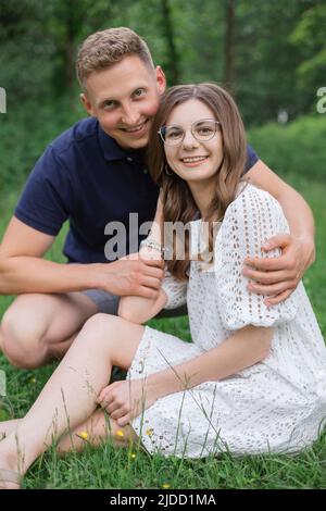 Giovane coppia affettuosa seduta sull'erba nel parco, guardando l'un l'altro. Giovane uomo e donna che si godono giorno d'estate. Gente felice nell'amore, luna di miele Foto Stock