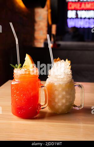 Limonata di fragole rosse con soda, ghiaccio, provetta da cocktail e fetta d'arancia in vetro con manico. Cocktail Pina Colada con ghiaccio, tubetto da cocktail e ananas Foto Stock