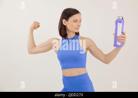 Ritratto di giovane donna sportiva con capelli scuri tenendo e guardando la bottiglia di plastica viola di acqua, mostrando i muscoli. Foto Stock