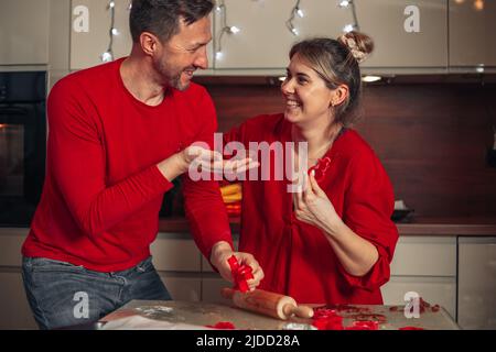 Giovane donna carina e uomo vestito di vestiti rossi ridono allegro mentre si cuocono i biscotti. Momenti di Natale, felice giovane coppia. Casa cucina accogliente. Foto Stock