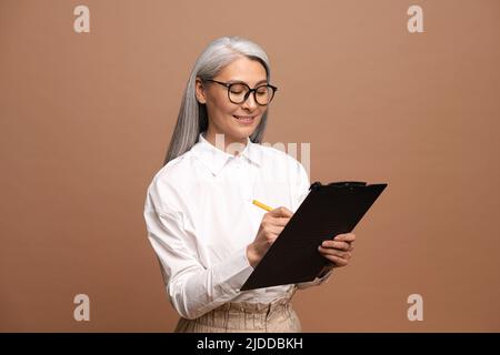 Senior fiducioso business donna scrittura negli appunti. Dipendente femminile maturo in abbigliamento formale isolato su sfondo beige Foto Stock