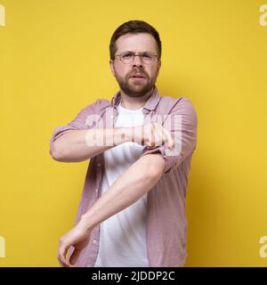 Calmo uomo caucasico in occhiali, rotola le maniche della camicia e coetanei in lontananza. Sfondo giallo. Foto Stock