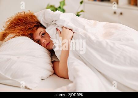Ragazza pigra rossa nel letto. Una ragazza pigra zenzero che si trova nel suo letto al mattino, coprendo la testa con un lenzuolo, e non vogliono alzarsi. Foto Stock